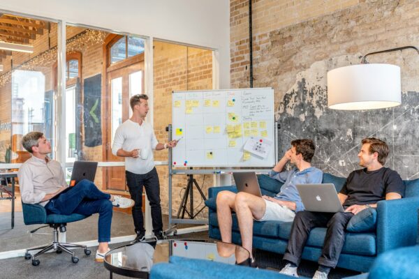 Un grupo de empleados discutiendo las mejores formas de posicionar su marca en una oficina moderna. fuente de imagen: https://unsplash.com/photos/three-men-sitting-while-using-laptops-and-watching-man-beside-whiteboard-wD1LRb9OeEo